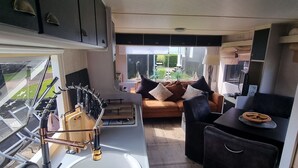 Een compact keuken met een spoelbak, fornuis en diverse keukengerei. Er is een eethoek met een tafel en stoelen, en uitzicht op de omgeving door een groot raam.