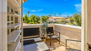 A balcony with a table and chairs, a potted plant, and a view of a residential area with houses and palm trees.