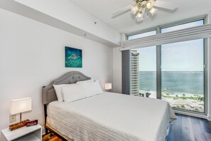 Une chambre à coucher comprenant un lit, une table de chevet, une horloge numérique, un ventilateur de plafond et une grande fenêtre donnant sur la mer et un immeuble.