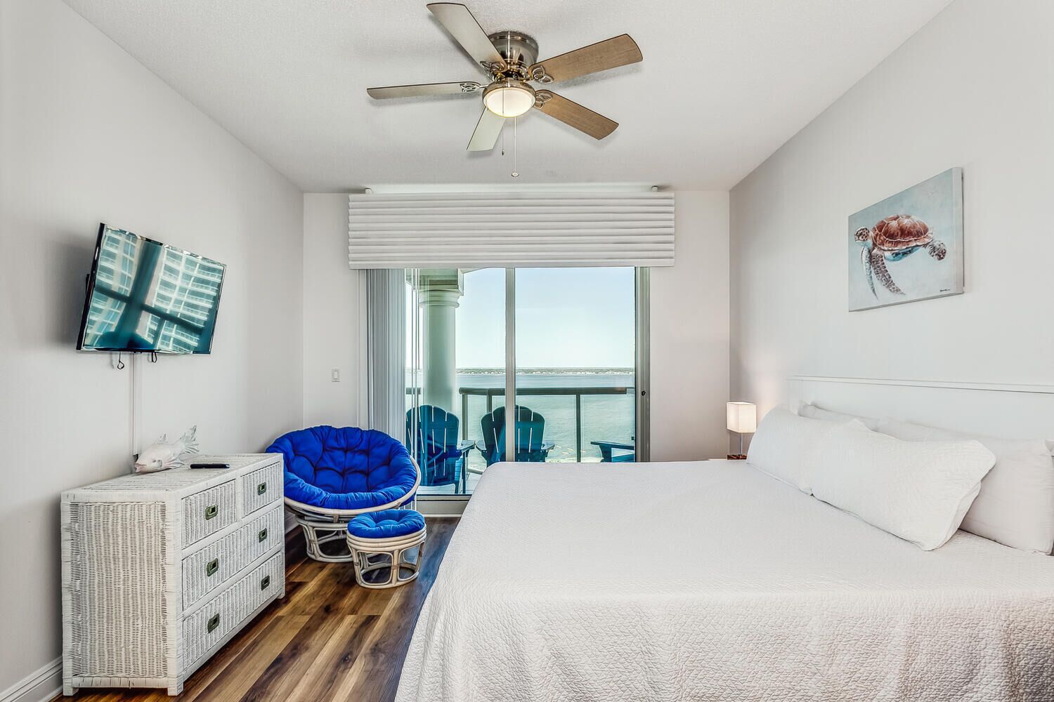 A hotel room with a bed, a ceiling fan, a television, a wicker dresser, a blue chair, and a view of the sea.
