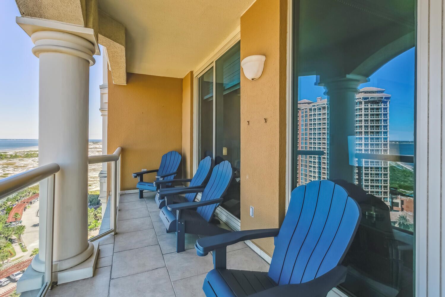 A balcony with four blue Adirondack chairs, a white balustrade, and a view of a cityscape and beach.