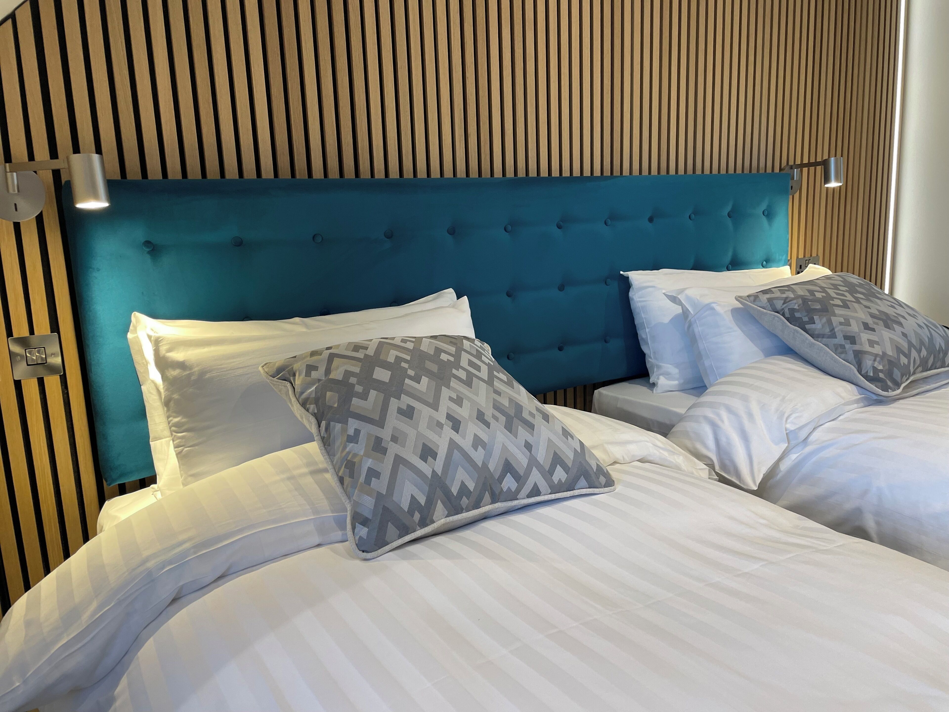 Una habitación de hotel con un cabecero tapizado en color verde azulado, ropa de cama blanca y almohadas estampadas.