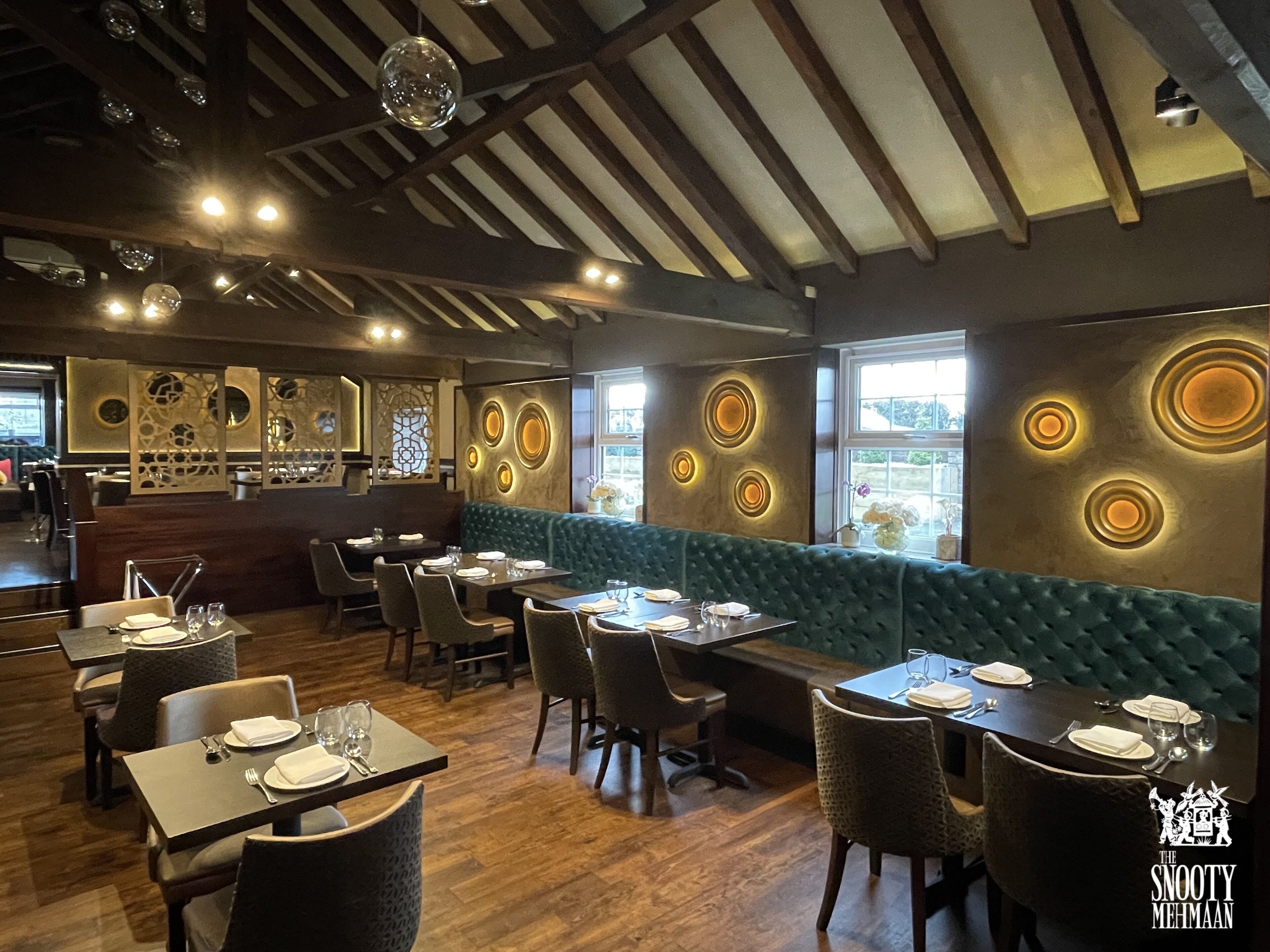 A restaurant interior with wooden flooring, green upholstered booths, and circular wall decorations. Tables are set with white tablecloths and silverware.