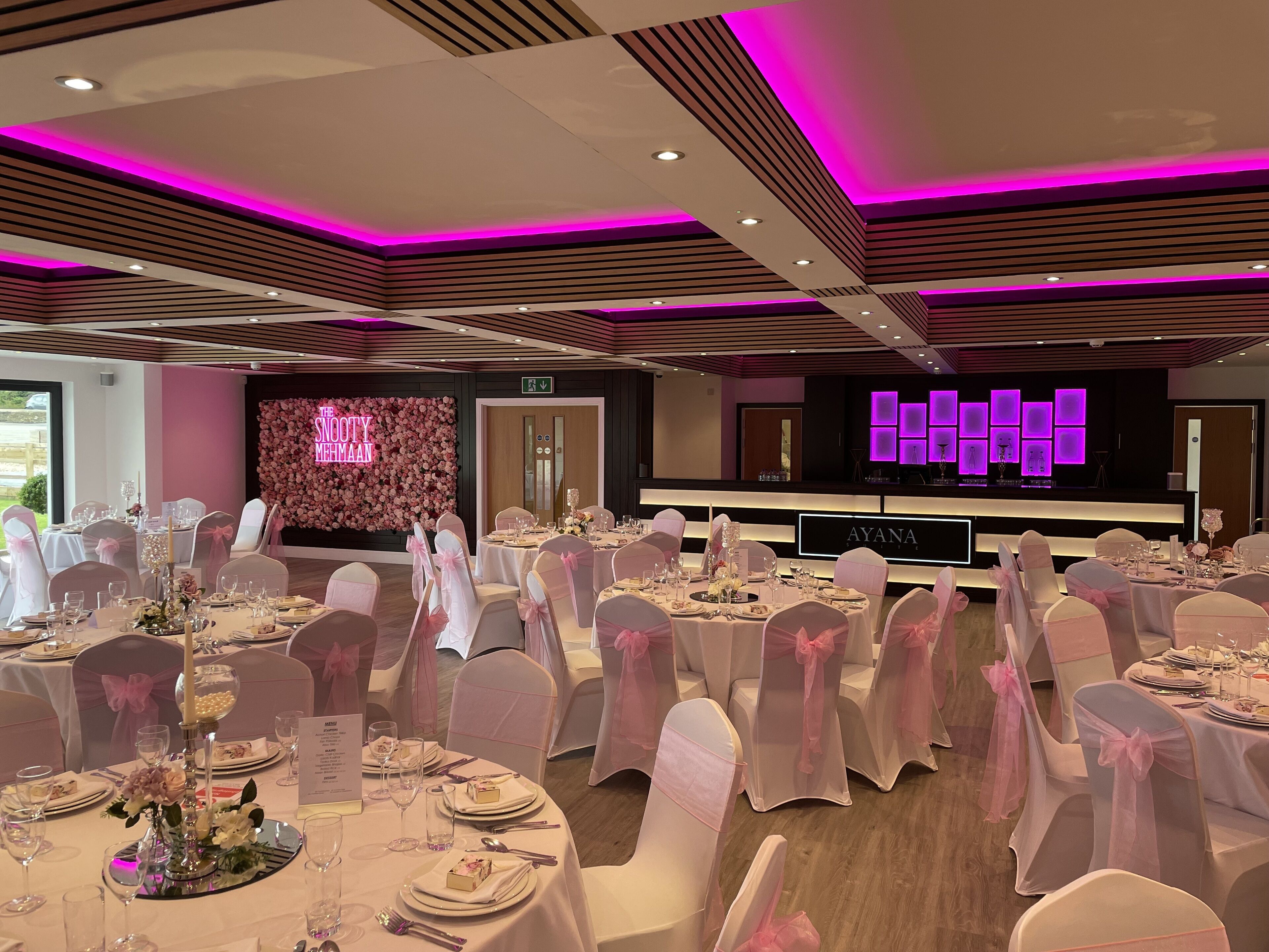 A banquet hall with round tables set for a formal event, featuring white tablecloths and pink chair covers.