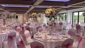 A banquet hall with round tables, white chairs with pink sashes, and floral centerpieces. - The Boutique Hotel by The Snooty Mehmaan (Faringdon)