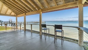 Ett takförsett utomhusområde med sittplatser, betonggolv och träbjälkar, med utsikt över en strandpromenad med en brygga och stolar.