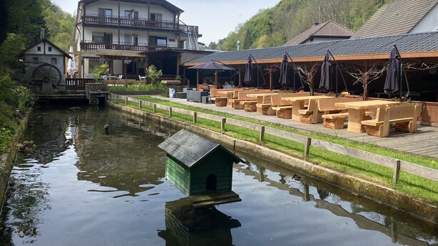 A riverside restaurant with outdoor seating, a wooden house, and a large building with a balcony. - Theis-Mühle (Biersdorf am See)
