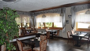 A dining area with wooden tables and chairs, large windows with curtains, and potted plants. - Theis-Mühle (Biersdorf am See)