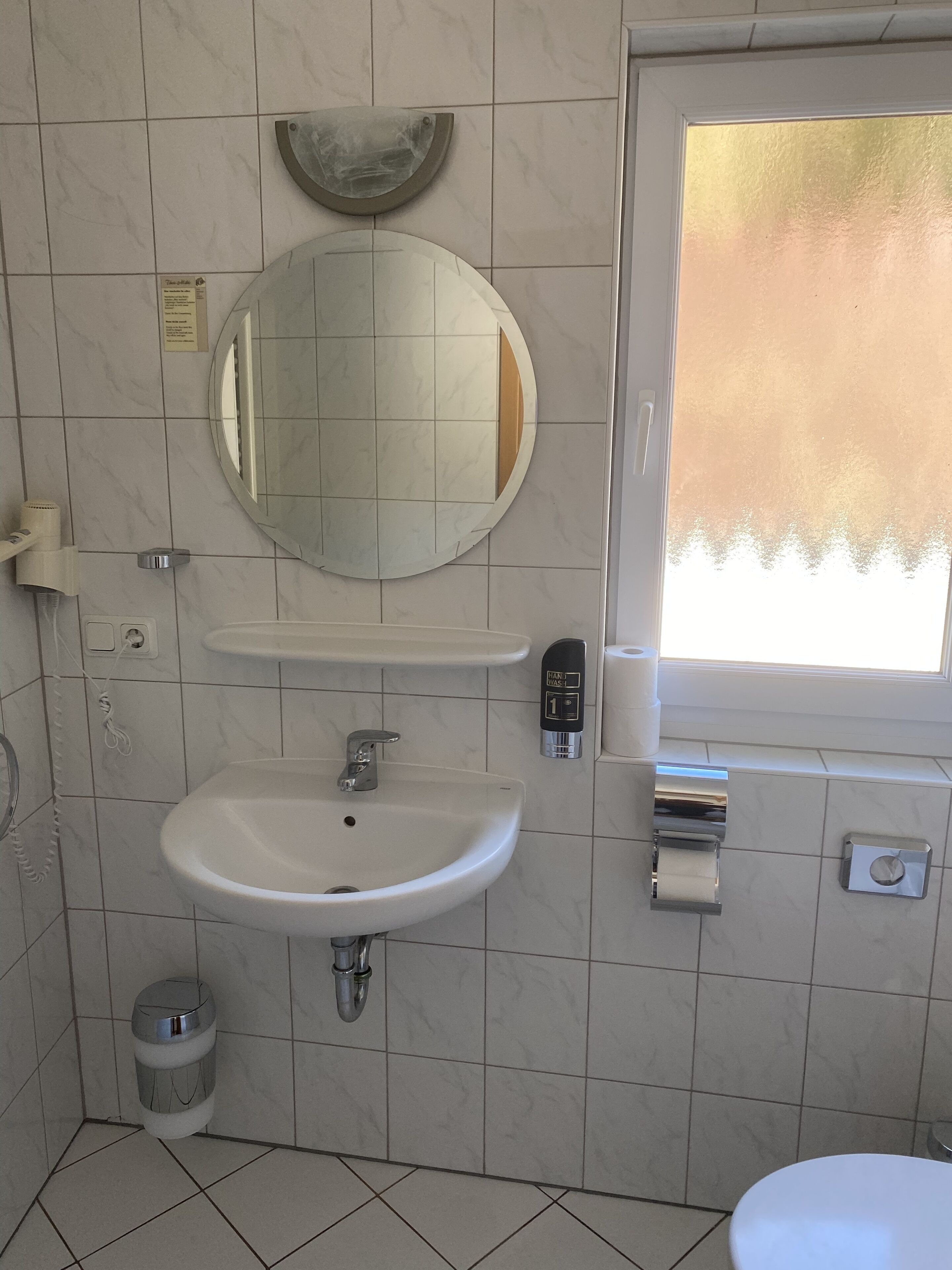 Un baño con lavamanos, espejo y tocador blanco, un inodoro y un cubo de basura.