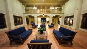 Un hall d’entrée spacieux avec de hauts plafonds, un espace de détente central avec des canapés et une table basse, et un balcon donnant sur l’intérieur.