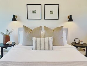 Een net opgemaakt bed met twee kussens, een nachtkastje met een klok en een kaars, en twee ingelijste foto's aan de muur.