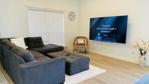A modern living room with a sectional sofa, a small armchair, a flat-screen TV mounted on the wall, and a decorative vase on a side table. - Luxurious home /Pool/GameR/BBQ/kingbed/legacyW/Frisco (plano)