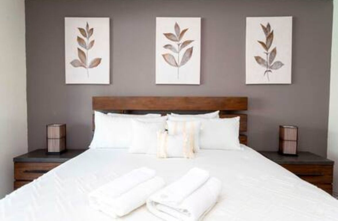 Ein ordentlich bezogenes Bett mit weißen Leinen und aufgerollten Handtüchern, ein Holz-Kopfbrett und drei an der Wand befestigte Kunstwerke mit Blättermotiv.