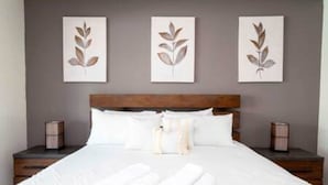 Una cama bien hecha con sábanas blancas y toallas enrolladas, un cabecero de madera y tres obras de arte con motivos de hojas.