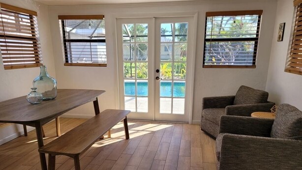 A room with a wooden floor, a bench, a table, a sofa, and a sliding glass door leading to a pool.