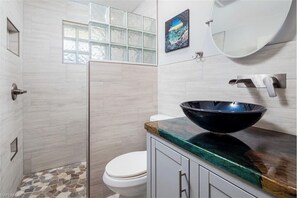 A bathroom with a glass shower enclosure, a white toilet, a large black vessel sink, and a painting above the mirror. - Home with pool - walk or bike to beach! (Naples)