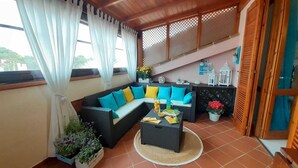 Un espacio exterior acogedor con un sofá negro y cojines azules, una mesa redonda con frutas y bebidas, y vistas al exterior a través de amplias ventanas.