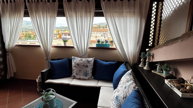 Una habitación acogedora con techo de madera, un sofá con cojines azules, una mesita con una planta y vistas a edificios a través de la ventana.