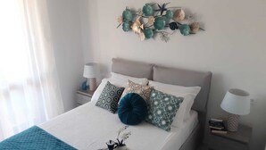 Una cama bien hecha con almohadones estampados, un adorno en la pared, mesitas de noche con lámparas y una bandeja con tetera y tazas.