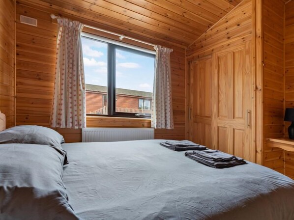 A wooden cabin room with a bed, a window with curtains, and a view of a building outside.