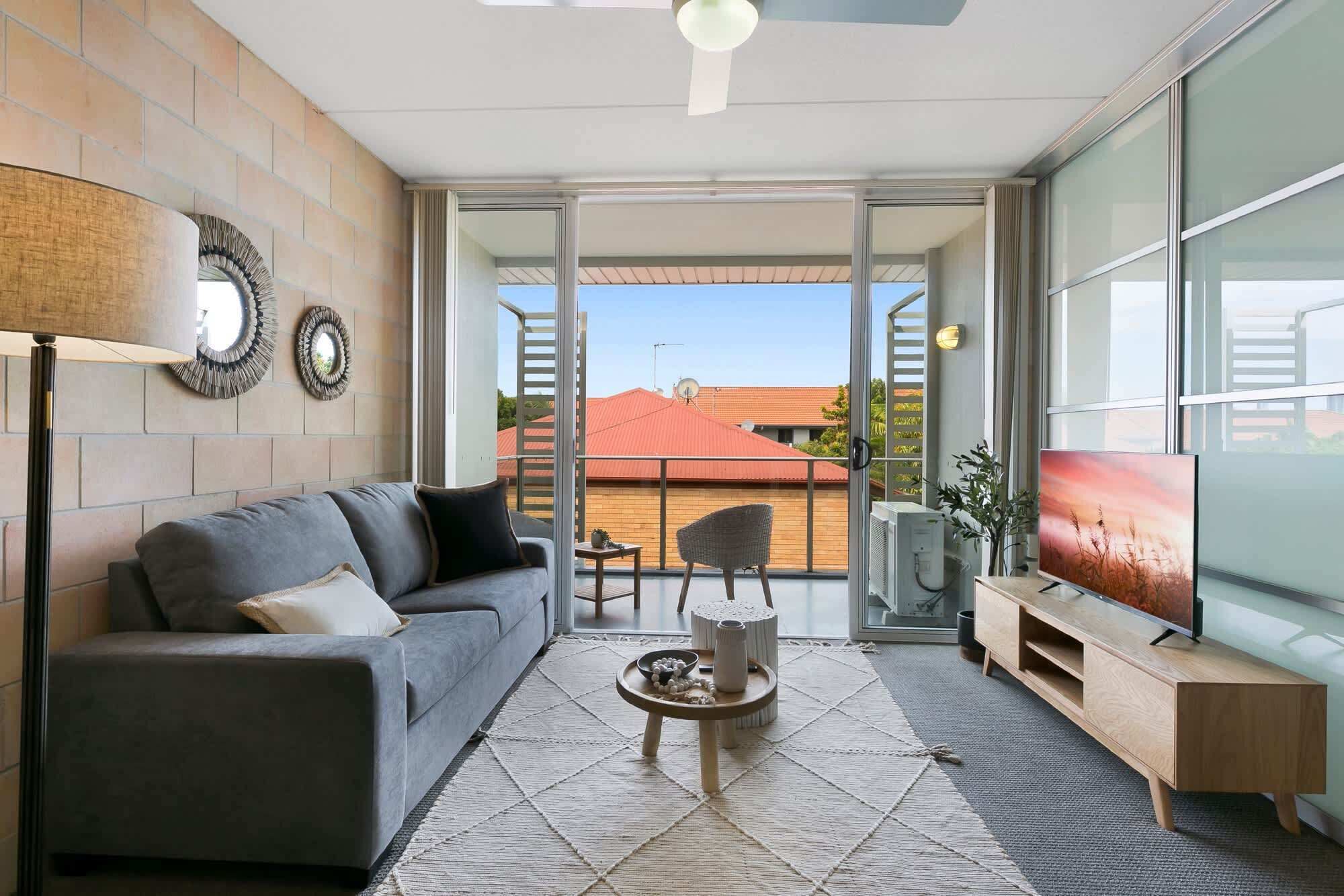 A modern living room with a grey sofa, a coffee table, a TV, and a balcony with a view of a red-roofed building.