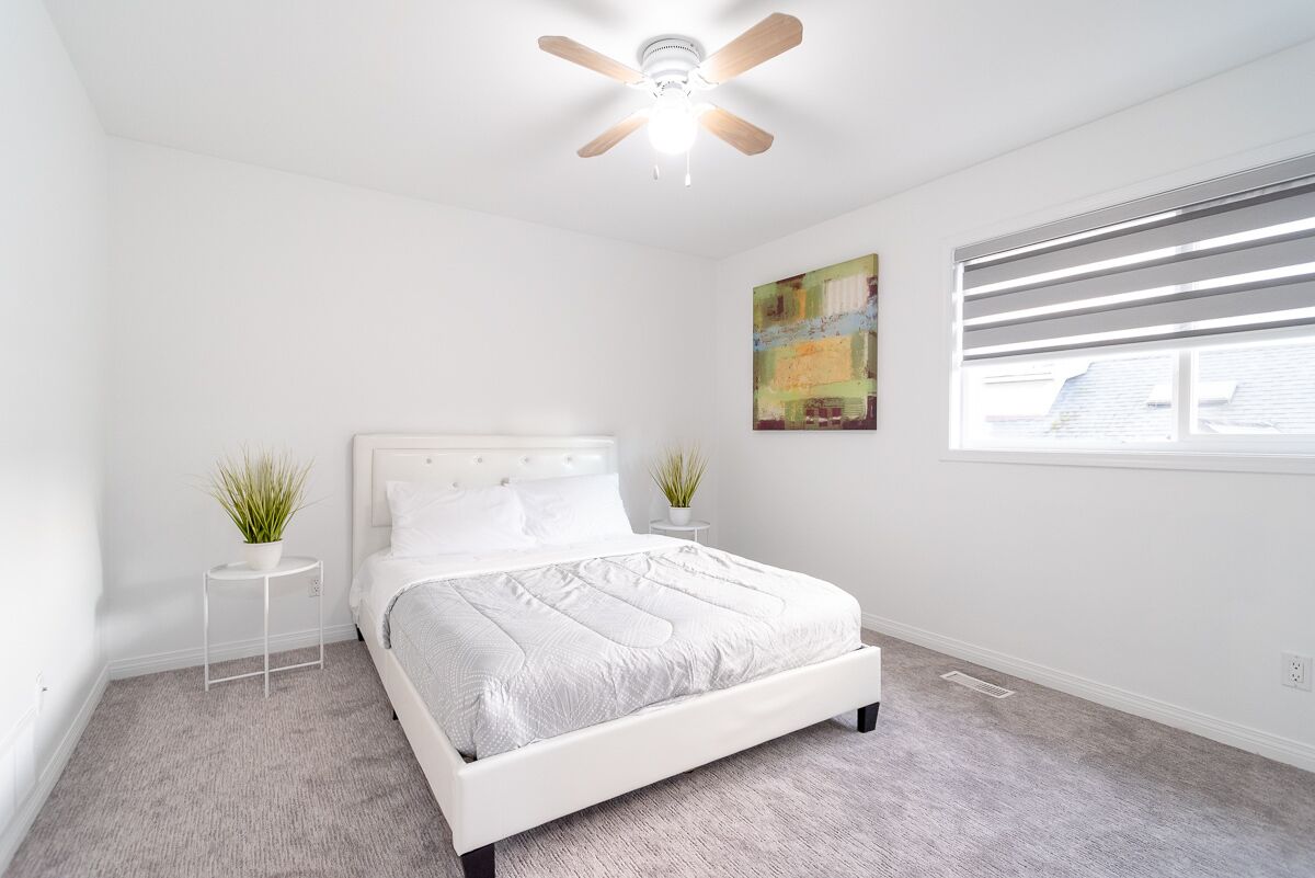 A bedroom with a white bed, a ceiling fan, a window with blinds, and a small side table with a plant.