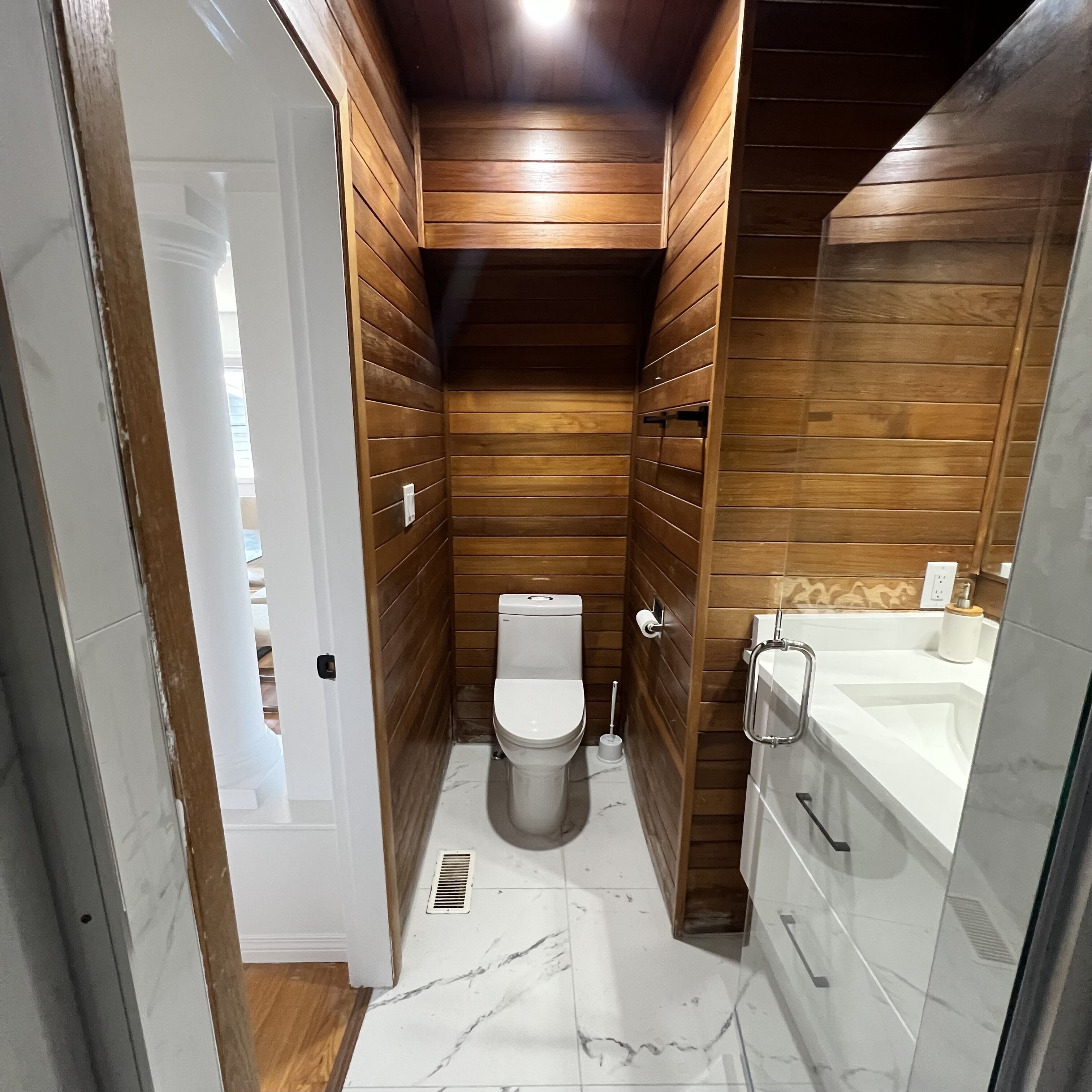 A compact bathroom with wooden paneling, a toilet, a sink, and a shower area.