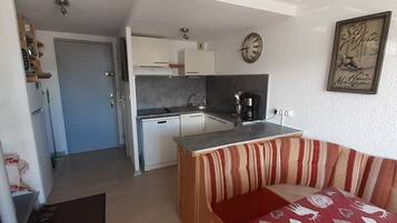 A compact kitchen with white cabinets, a sink, and a coffee maker. There is a striped sofa in the foreground and a clock on the wall.