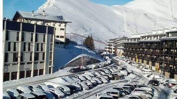 A snow-covered parking lot with cars and buildings, including a hotel with balconies and a mountain in the background.