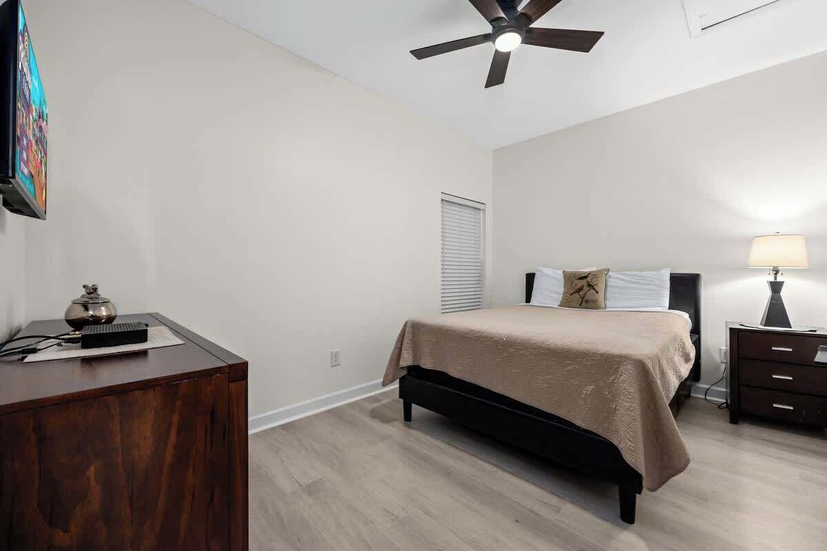 A bedroom with a bed, a desk, a ceiling fan, a lamp, and a window with blinds.