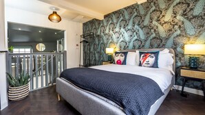 A modern bedroom with a large bed, a decorative wall, a small table with a lamp, and a potted plant. - The Book Club, York. (York)