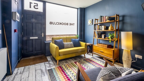 A modern living room with a yellow sofa, a bookshelf, and a large window with a view. - The Book Club, York. (York)