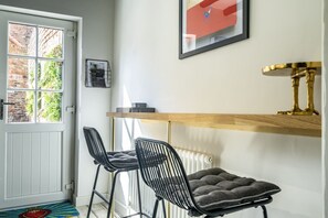 A compact room with a white door, a radiator, a small table with a gold base, two black chairs, and a cushioned bench.