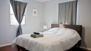 A bedroom with a large bed, two windows with curtains, a bedside table with a lamp, and a framed picture on the wall.