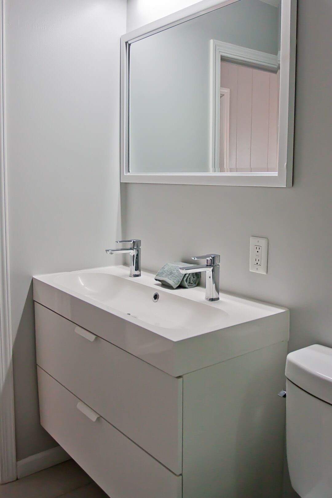 Een badkamer met een wit wastafelmeubel, twee kraanwerkstukken, een spiegel en een toilet.