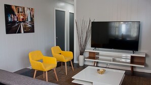 A modern living room with a large flat-screen TV, two yellow chairs, a white coffee table, and a painting on the wall.