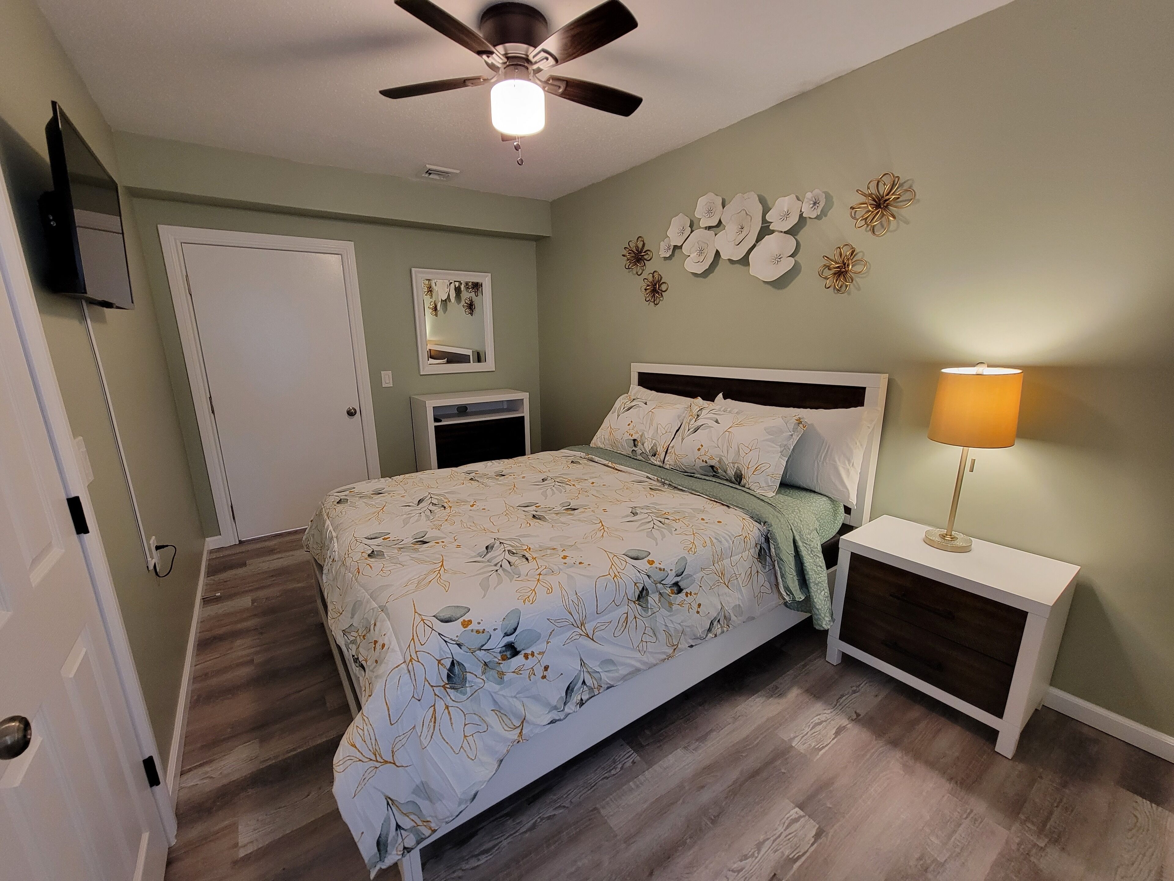 A bedroom with a bed, nightstands, a lamp, a ceiling fan, a TV, a door, and a wall with decorative items.