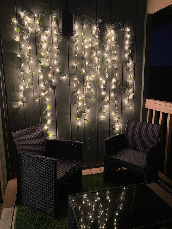 Un mur en bois foncé, décoré de guirlandes lumineuses, et un miroir qui reflète la lumière. On y trouve deux fauteuils en osier et une petite table avec un plateau en verre.