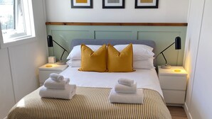 Un lit bien fait, recouvert d’une couvre-lit à motifs gris et jaunes, avec deux coussins de couleur moutarde et deux serviettes blanches pliées sur le côté. Au mur, au-dessus du lit, on peut voir trois tableaux encadrés. De chaque côté du lit, il y a une table de chevet avec une lampe, et une fenêtre se trouve sur le côté gauche.
