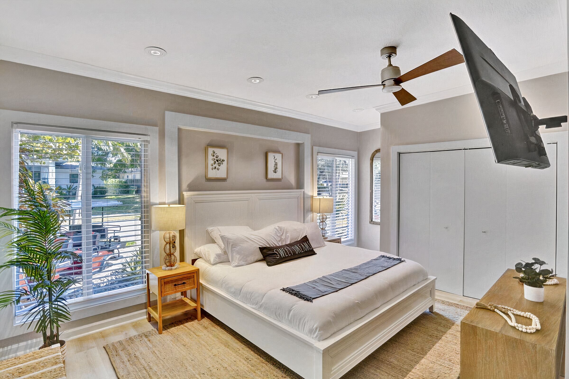 A bedroom with a large bed, a ceiling fan, a TV mounted on the wall, and a window with blinds.
