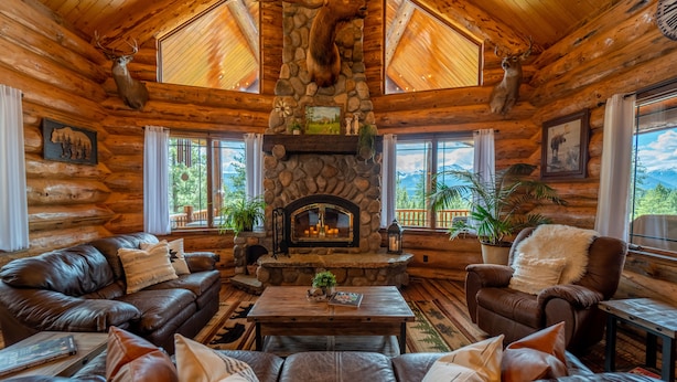 L’intérieur d’une cabane en rondins, avec une cheminée en pierre, des meubles en cuir et une vue sur les montagnes.