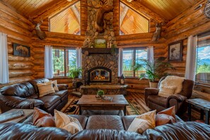 A log cabin interior with a stone fireplace, leather furniture, and a mountain view. - Montana Haven - A Peaceful Mountain Retreat (Libby)