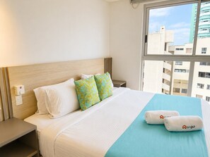 Habitación de hotel con cama, almohadas, mesita de noche, ventana con vistas a edificios y panel de control en la pared.