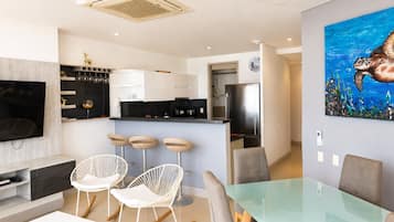Een moderne woonkamer met een televisie, witte schommelstoelen, een glazen eettafel en een keuken op de achtergrond.