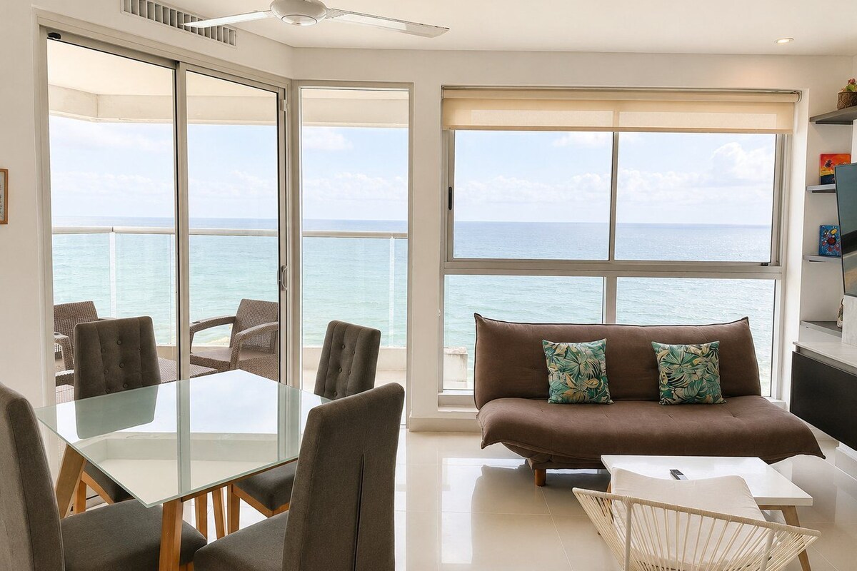 A modern living room with a glass dining table, two sofas, a white coffee table, and a view of the ocean through large windows.
