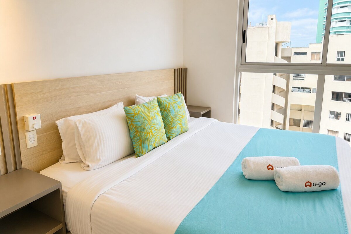 A hotel room with a bed, pillows, a nightstand, a window with a view of buildings, and a control panel on the wall.