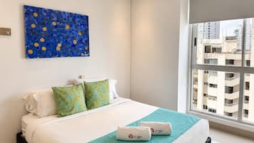Een hotelkamer met een bed, kussens, een blauw kunstwerk aan de muur en een raam met uitzicht op de stad.