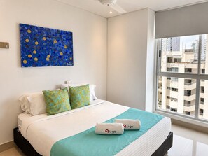 A hotel room with a bed, pillows, a blue artwork on the wall, and a window with a city view. - U-GO Edificio Murano Elite, Bocagrande (Cartagena)