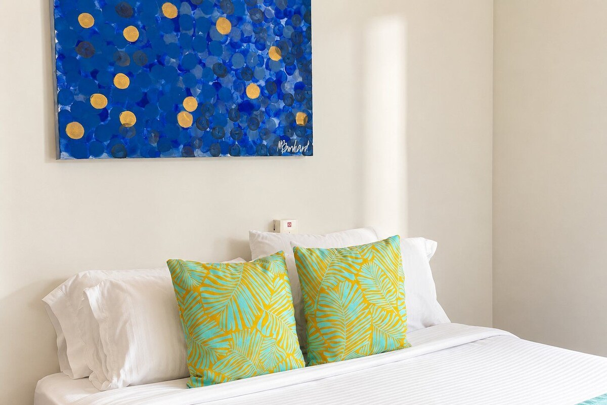 A hotel room with a bed, white bedding, two decorative pillows with a leaf pattern, a wall-mounted artwork with blue and yellow circles, and a white nightstand.