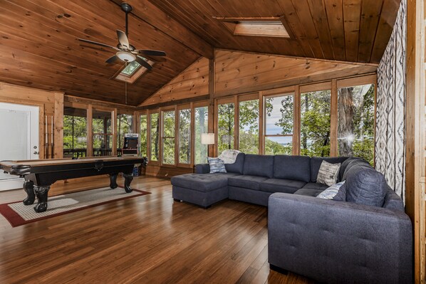 A spacious room with a pool table, a sectional sofa, and large windows offering a view of the outdoors.